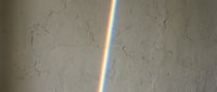 Rainbow light prism reflection on textured beige concrete wall, colorful spectrum streak creating vibrant diagonal line across rough plaster surface.