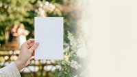 A woman's hand holding a blank white card outdoors with blooming flowers and green foliage in soft focus background, suitable for greeting cards, invitations, and promotional messaging.