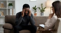 African American man experiencing stress during therapy session with female counselor in modern office setting discussing mental health concerns.