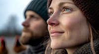 Young Caucasian woman in knit beanie smiling while looking up at winter sky, with bearded man in background. Natural outdoor lighting creates warm atmosphere.
