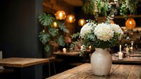 Cozy restaurant interior with white flower bouquet in ceramic vase on wooden table, warm ambient lighting and greenery creating inviting dining atmosphere.