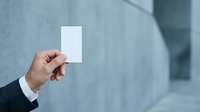 Man in business suit holding blank white card against modern building facade for corporate branding and professional networking purposes.