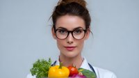Young female healthcare professional in white coat and glasses holding fresh vegetables and fruits, promoting healthy eating and nutrition counseling concept.