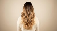 Young woman with long wavy blonde ombre hair from behind wearing white sweater against beige background for hair care and beauty concepts.