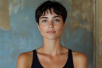 Young woman with short dark pixie haircut and natural makeup wearing black tank top against textured blue wall background, looking directly at camera with confident expression.