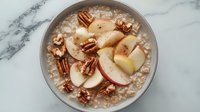 Creamy oatmeal bowl topped with fresh sliced red apples and crunchy pecan nuts on marble surface, healthy breakfast meal rich in fiber and protein.