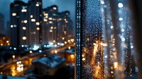 Raindrops on window glass at night with blurred city lights and urban buildings in background creating atmospheric moody scene with bokeh effect and reflections.