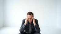 Stressed middle-aged Caucasian businessman in dark suit holding head in hands showing work pressure, mental fatigue and corporate burnout at office desk.