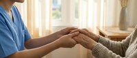 Healthcare worker in blue scrubs holding hands with elderly patient providing comfort and compassionate care in bright medical facility room.