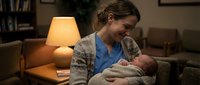 Young Caucasian mother in cozy cardigan holding sleeping newborn baby in warm living room with soft lamp light creating intimate bonding moment at home.