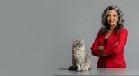 Middle-aged woman with gray wavy hair and light skin tone, wearing a bright red blazer, smiling confidently while posing next to a fluffy gray tabby cat against a neutral gray background.