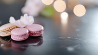 Delicate French macarons in soft pink and white tones on dark reflective surface with blurred bokeh lights creating romantic atmosphere for food photography.