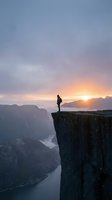 Silhouetted hiker standing on rocky cliff edge overlooking misty canyon valley at golden sunrise, contemplating vast landscape with mountains and river below.