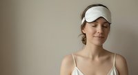 Young woman wearing white sleep mask relaxing with peaceful expression in soft natural lighting for wellness and self-care routines.