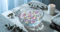 Spa wellness setup with glass bowl filled with water, floating pink rose petals and white daisies, eucalyptus leaves, candle and towels for relaxation.