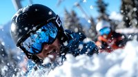 Young male skier in protective helmet and blue mirrored goggles spraying fresh powder snow while skiing downhill, with another skier visible in background on sunny day.