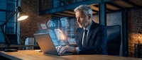 Professional mature businessman working on laptop with holographic digital interface displays in modern industrial office space for technology presentations.