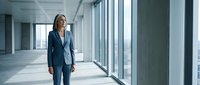 Confident middle-aged Caucasian businesswoman in navy blazer walking through modern office corridor with floor-to-ceiling windows, representing corporate leadership and professional success.