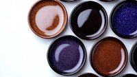Shimmering cosmetic pigments and glitter powders in round containers on white background, featuring metallic copper, black, purple and blue tones.