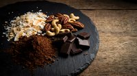 Cocoa powder, dark chocolate pieces and mixed nuts arranged on black slate board for baking and confectionery preparation on rustic wooden background.