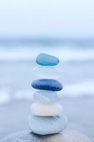 Balanced stack of smooth sea glass stones in blue and white tones on beach with ocean waves background for meditation and wellness concepts.