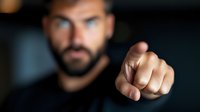 Dramatic close up of pointing finger gesture against dark background, selective focus on hand while bearded man stands blurred in background.