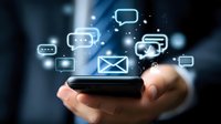 Hand holding smartphone with glowing message icons and chat bubbles floating above in dark blue background, representing digital communication and mobile messaging.