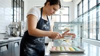 Professional female baker decorating colorful macarons in modern commercial kitchen wearing apron. Pastry chef preparing French desserts for bakery display.