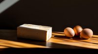 Fresh farm eggs and butter block on wooden cutting board with dramatic morning sunlight and shadows creating minimalist culinary composition.