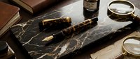 Luxury fountain pen with tortoiseshell barrel resting on black marble surface alongside vintage books, magnifying glass and inkwell for writing, journaling and stationery design projects.