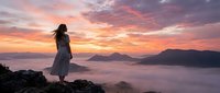 Woman in flowing dress standing on rocky mountain peak overlooking misty valley at sunrise, golden and pink sky with clouds, contemplative travel and nature scene for editorial and lifestyle use.