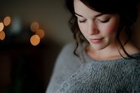 Young Caucasian woman in gray knit sweater smiling gently with eyes closed, soft bokeh lights in dark background creating intimate atmospheric portrait.