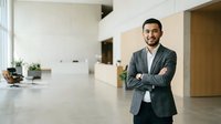 Confident male professional with dark hair and beard, South Asian ethnicity, wearing gray blazer and white shirt, standing with crossed arms in modern office lobby with minimalist design and natural lighting.