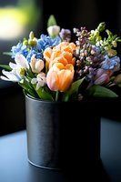 Fresh spring bouquet with peach tulips, blue hyacinths, and delicate buds arranged in black metal container against dark background. Natural lighting highlights floral details.
