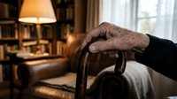 Elderly hands resting on wooden walking cane in cozy home library setting with warm lighting and bookshelves creating peaceful retirement atmosphere.