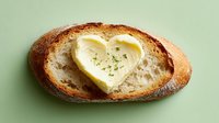 Artisan bread slice with heart-shaped butter pat and herbs on mint green background, perfect for food blogs, restaurant menus and culinary content.