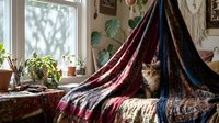 Cozy bohemian living room interior with tabby cat sitting on colorful patterned blankets near sunny window with houseplants and macrame wall decor.