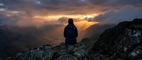 Person sitting on mountain rock overlooking dramatic sunset sky with golden clouds and valley below, contemplative outdoor adventure scene for travel and lifestyle use.