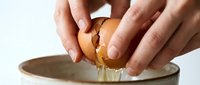 Caucasian woman's hands cracking a fresh brown egg over a ceramic bowl, with yolk and whites dripping down, captured in natural light.