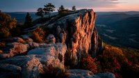 Dramatic rocky cliff formation glowing in warm sunset light overlooking mountain valley landscape with autumn foliage and evergreen trees creating scenic vista.