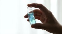 Hand holding small glass vial with blue liquid sample for laboratory analysis, research or medical testing in bright clinical environment.