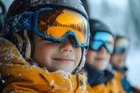Young skier in bright orange winter jacket and protective goggles smiling during winter sports activity, with other skiers in background on snowy day.