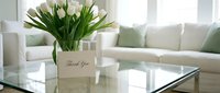 White tulips in a glass vase with a thank you card on a coffee table in a bright, elegant living room interior.