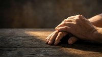 Elderly hands clasped together in prayer or meditation on wooden surface with warm lighting creating peaceful spiritual atmosphere for faith concepts.