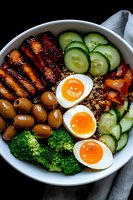 Healthy protein bowl with grilled chicken, soft boiled eggs, quinoa, fresh vegetables, broccoli, cucumber slices and green olives served in white bowl on dark background.