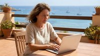Middle-aged woman with shoulder-length brown hair working on laptop at wooden table on coastal terrace overlooking Mediterranean sea, remote work and vacation lifestyle.