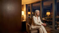 Confident mature businesswoman in elegant beige suit sitting in luxury hotel room with panoramic city night view through floor-to-ceiling windows.