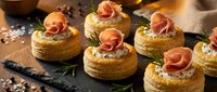 Elegant puff pastry appetizers topped with prosciutto roses and herbs on rustic slate board for gourmet entertaining and holiday party catering.