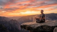 Young man meditating on mountain cliff edge during golden sunset with dramatic orange sky and canyon landscape for wellness and mindfulness concepts.