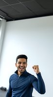 Young man with dark hair and olive complexion wearing navy blue zip-up athletic jacket, smiling broadly with fist raised in celebration gesture inside modern office space with white walls and dark ceiling tiles.